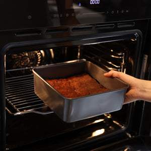 A hand is placing a Zyliss Non-Stick Base Square Pan 20cm/8", filled with freshly baked brownies, into or out of a modern black oven.