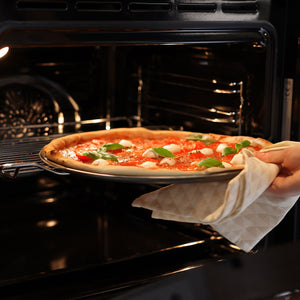 A hand with an oven mitt places a pizza topped with tomato sauce, mozzarella, and basil onto the Zyliss Non-Stick Pizza Tray, ensuring perfectly crispy bases in a hot oven.