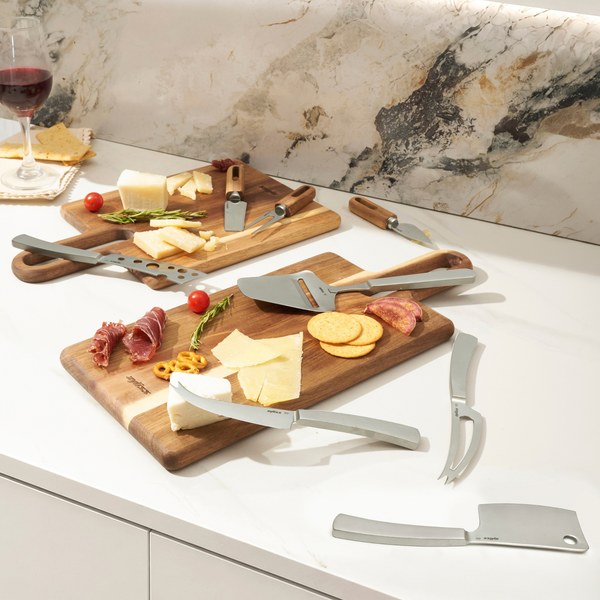 The Zyliss Signature 3 Piece Cheese Knife & Acacia Serving Board Set displays assorted cheeses, crackers, and meats on a white marble countertop, with a glass of red wine and napkin in the background. The scene is bright and modern.