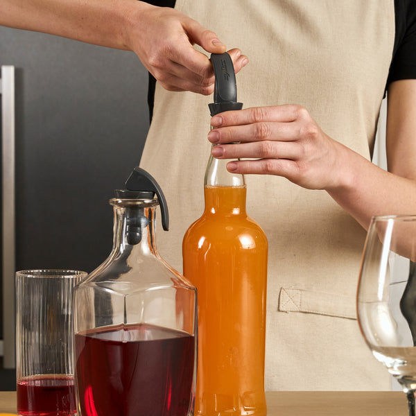 A person in a beige apron opens an orange bottle with a bottle opener beside a glass pitcher of red liquid, a glass, and an empty wine glass on wood—ideal for keeping drinks fresh with the Zyliss Bottle Stopper 2 Piece Set.