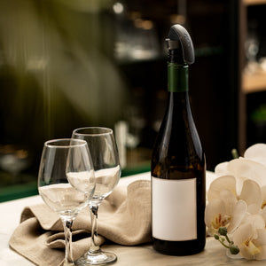 A wine bottle with a blank label and a Zyliss Bottle Stopper 2 Piece Set sits on a table near two empty glasses, a beige cloth, and white orchids. The softly blurred background enhances the elegant storage solution.