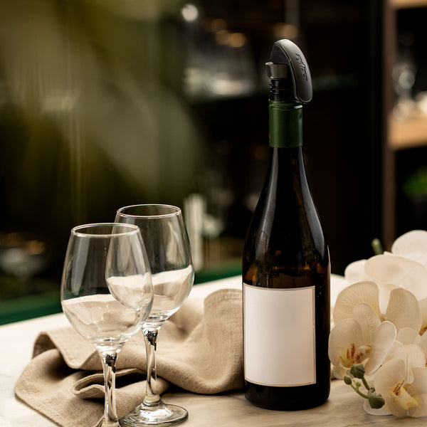 A wine bottle with a blank label and a Zyliss Bottle Stopper 2 Piece Set sits on a table near two empty glasses, a beige cloth, and white orchids. The softly blurred background enhances the elegant storage solution.