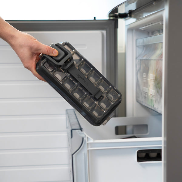 A hand places the Zyliss Press & Pull Ice Cube Tray with its lid into a partially open freezer drawer.