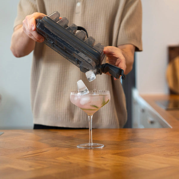 Someone pours ice cubes from the Zyliss Press & Pull Ice Cube Tray into a cocktail glass with a pink drink and lime slices, on a wooden kitchen counter.