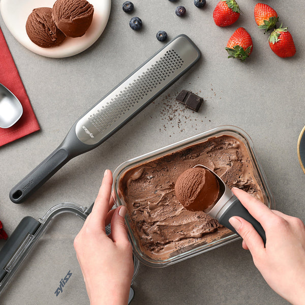 A person scoops chocolate ice cream from a rectangular Zyliss Glass Container. Nearby are a grater, chocolate pieces, blueberries, strawberries, an ice cream scoop, and a red napkin on a gray surface—ideal for easy meal prep or dessert storage!. #0.64L/0.67qt