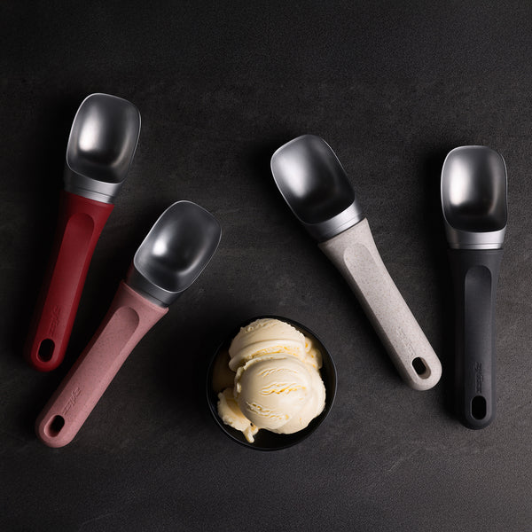 A bowl of vanilla ice cream rests on a dark surface, encircled by four Zyliss Ice Cream Scoops with eco-friendly red, pink, gray, and black handles—each neatly arranged and dishwasher-safe for easy cleaning.