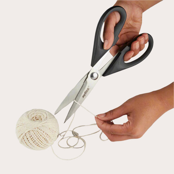 A pair of hands uses Zyliss Household Shears with stainless steel blades to cut white string from a ball of twine against a plain background. 