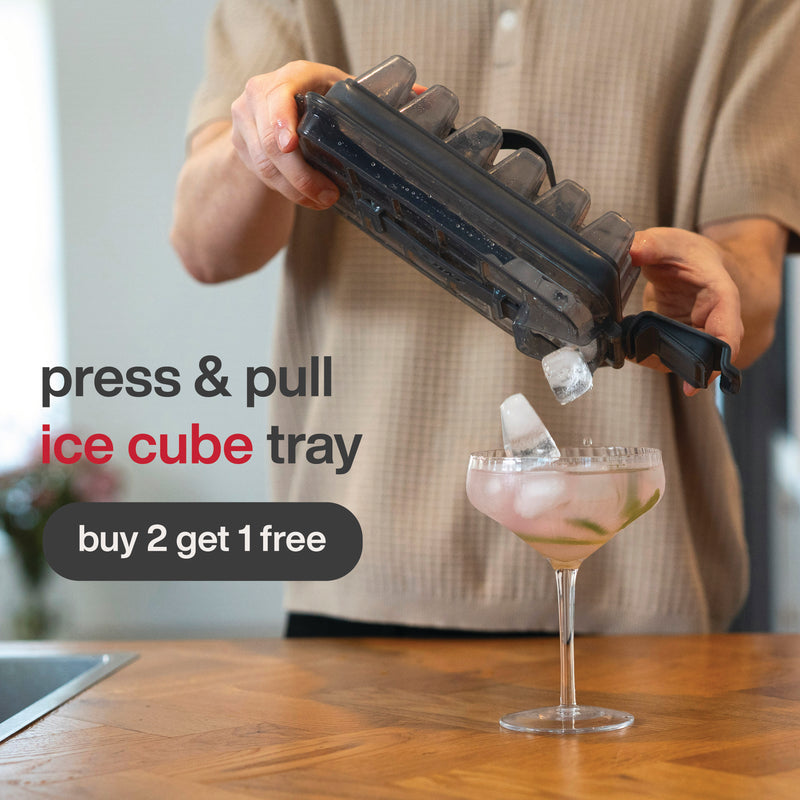 A person pours ice cubes from a black Zyliss Press & Pull Ice Cube Tray into a cocktail glass, with text reading, “Zyliss Press & Pull Ice Cube Tray – Buy 2 Get 1 Free.”.