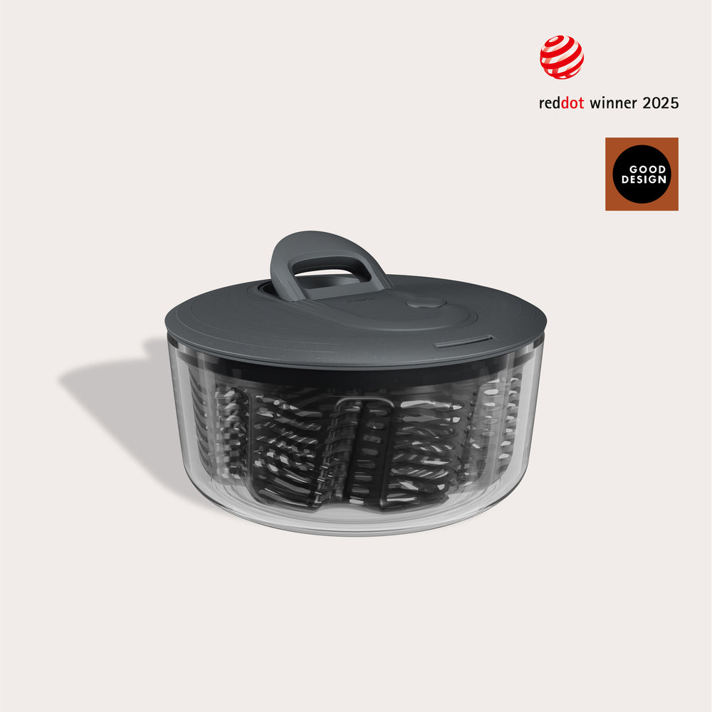 A Zyliss Easy Spin Salad Spinner with a clear bowl and dark gray lid is displayed on a light beige background. Red Dot Winner 2025 and Good Design logos appear in the top right corner.