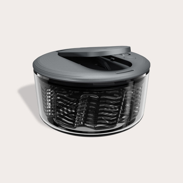 The Zyliss Swift Dry Salad Spinner features a transparent, round bowl with a dark gray lid and one-handed pump handle. Its slitted inner basket is visible through the clear outer bowl, shown on a light background.
