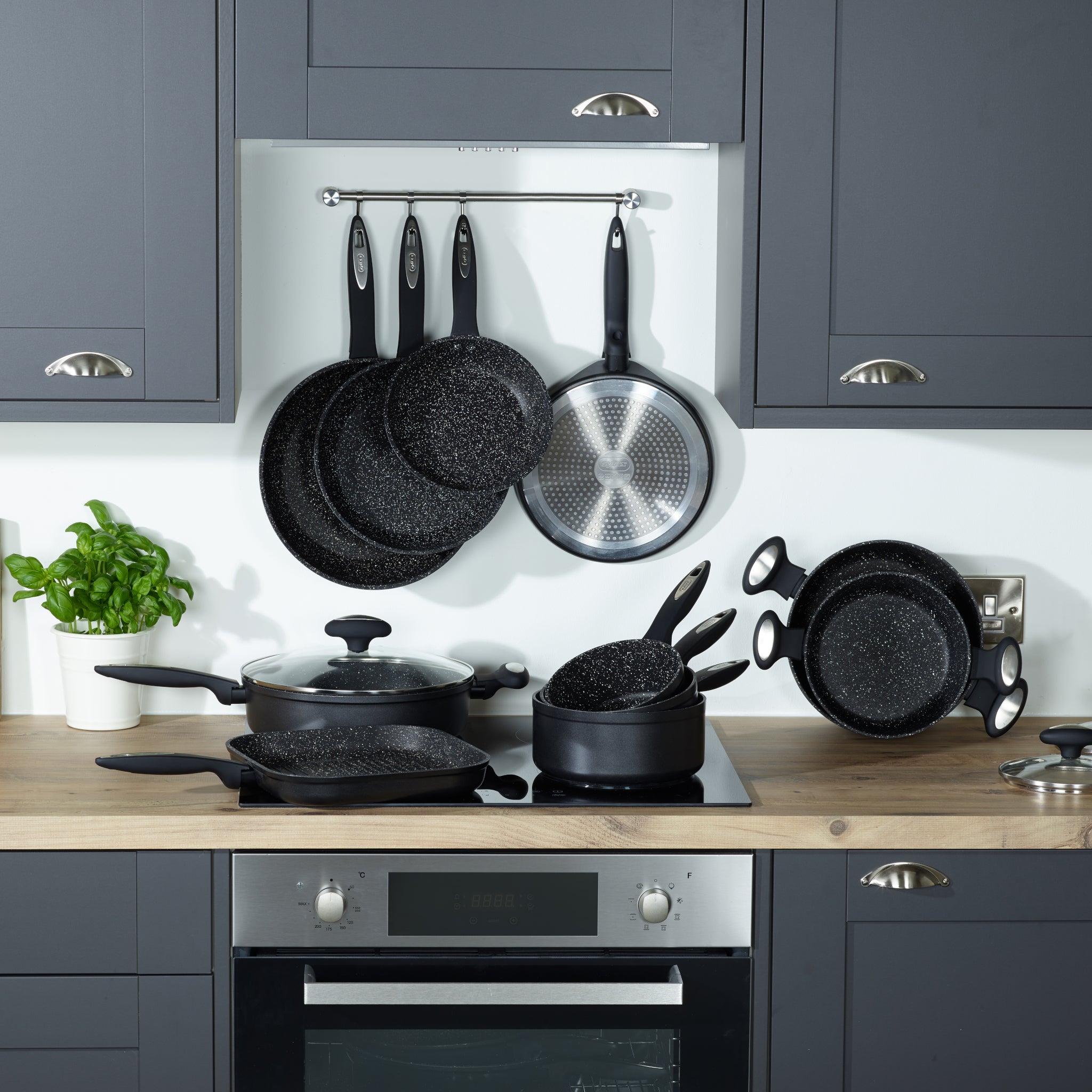A modern kitchen with dark cabinets, a built-in oven, and wooden countertop displays a set of black speckled pots and pans on the stove and hanging on the wall, with a basil plant on the counter.