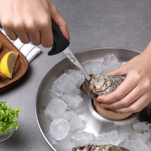 A person uses the Zyliss Kitchen Oyster Knife Shucker Paddy to open an oyster over a metal bowl with ice and more oysters. Nearby are a cutting board, lemon wedges, lettuce, and a towel on the table.