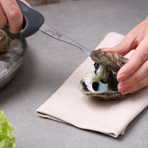 A person uses a Zyliss Kitchen Oyster Knife Shucker Paddy to open a fresh oyster on a beige napkin, with a bowl of ice and lettuce nearby on a gray surface.