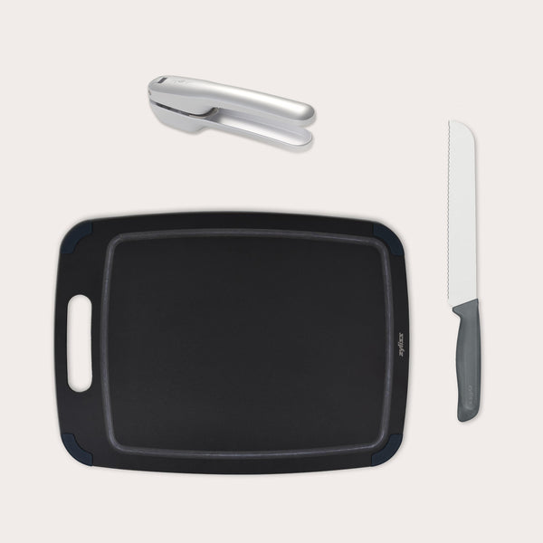The Zyliss Garlic Bread Making Kit features a black cutting board with handle, silver garlic press, and gray-handled serrated bread knife, ideal for perfect garlic bread, all arranged on a light background. #large