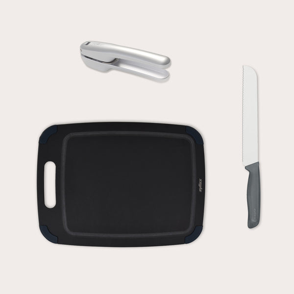The Zyliss Garlic Bread Making Kit features a black handled cutting board, a silver garlic press, and a gray-handled bread knife—everything you need for garlic bread, elegantly arranged on a light background. #medium