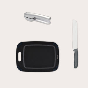 The Zyliss Garlic Bread Making Kit, featuring a metal garlic press, black cutting board with handle, and bread knife with gray handle, is neatly arranged on a light background—ideal for your kitchen. #small