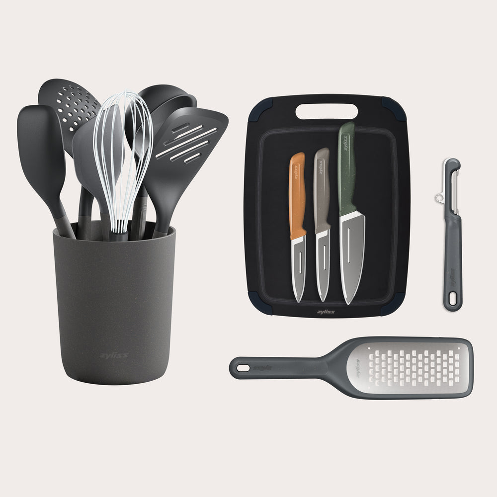 The Zyliss Kitchen Essentials Bundle features eco-friendly utensils with holder, three stainless steel knives on a cutting board, a veggie peeler, and handheld grater—all neatly arranged on a white background.