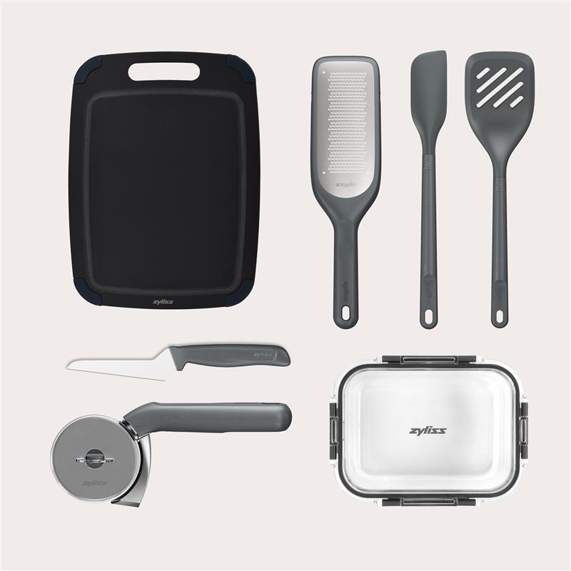 The Zyliss 7-Piece Kitchen Essentials Bundle includes a black cutting board, grater, silicone and slotted spatulas, pizza cutter, food container with transparent lid—all displayed on a light background.