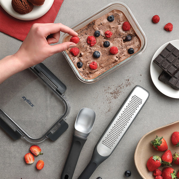 A hand decorates a container from the Zyliss Dessert Set with fresh berries. Beside it are a metal scoop, glass storage boxes, strawberries, chocolate pieces, and scattered berries on a gray countertop.