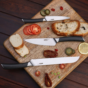 The Zyliss Comfort Pro Chef + Santoku Knife Set, featuring three kitchen knives with black handles and stainless steel blades, rests on a wooden cutting board surrounded by sliced bread, tomato, lemon, pickles, chili, and sun-dried tomatoes. The board is placed on a dark wooden surface.