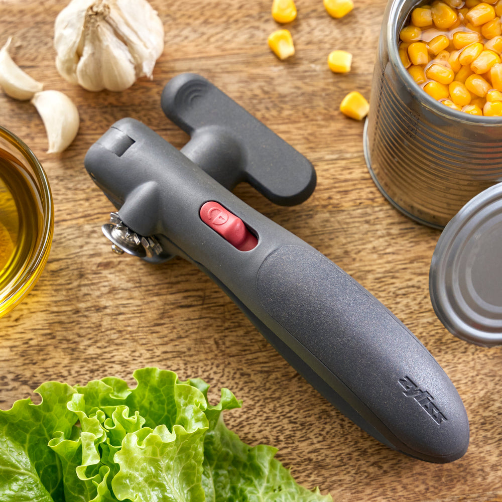 A dark gray ergonomic tool with a textured finish, featuring a small red button and a silver metal ring, shown on a solid white background.