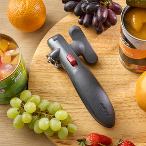 A black manual can opener with a red switch, displayed on a wooden board surrounded by fruit including grapes, strawberries, oranges, and canned fruit.