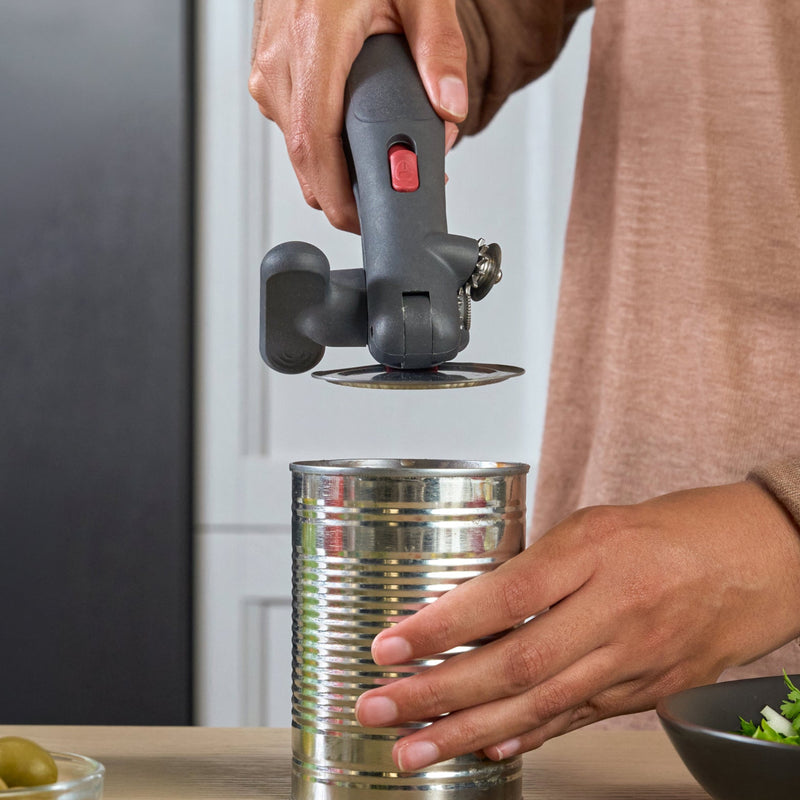 A person using a manual can opener to open a metal can on a kitchen counter, with canned olives, salad, and other kitchen items nearby.