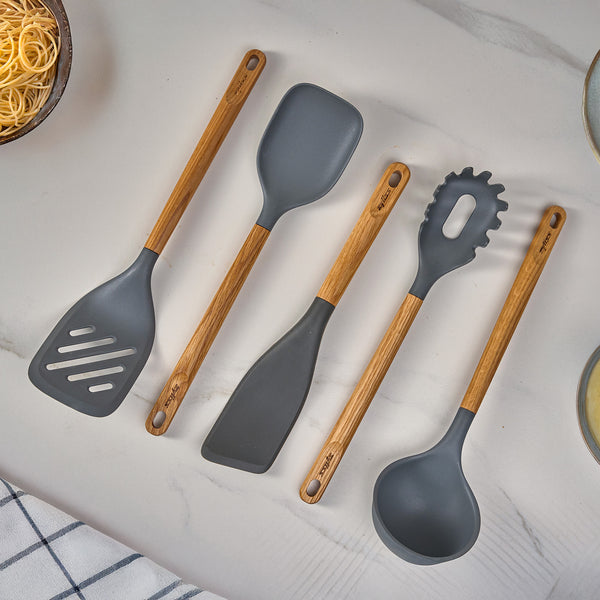 The Zyliss Oak Wood Utensil Set features four gray-headed tools—slotted spatula, scraper, spaghetti server, and ladle—displayed on a marble countertop beside plates, pasta, and a checkered towel.