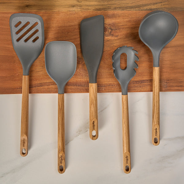 The Zyliss Oak Wood Utensil Set features five utensils—slotted spatula, spoon, scraper, pasta server, and ladle—with grey silicone heads and oak handles, displayed side by side on a wood and marble surface.
