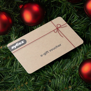A Zyliss e-Gift Voucher rests on green pine branches amid red Christmas ornaments. The tan voucher, featuring a festive red string graphic, makes a perfect holiday gift for premium kitchenware from Zyliss.