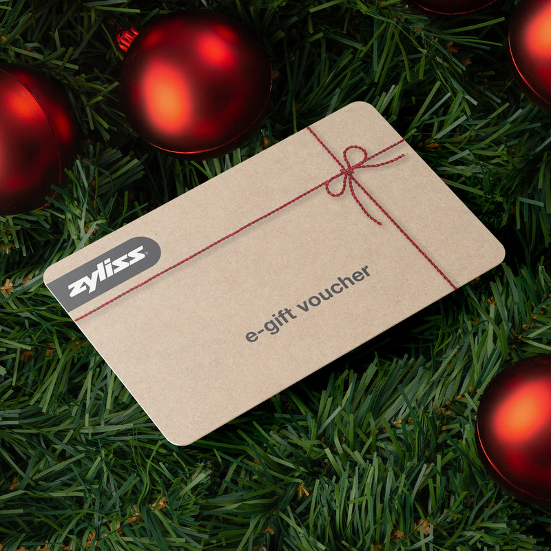 A Zyliss e-Gift Voucher rests on green pine branches amid red Christmas ornaments. The tan voucher, featuring a festive red string graphic, makes a perfect holiday gift for premium kitchenware from Zyliss.