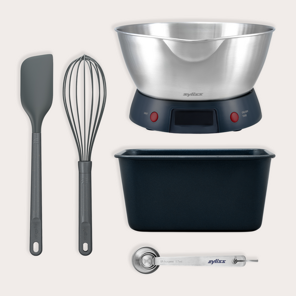 The Zyliss Banana Bread Baking Essentials Kit includes a metal mixing bowl, digital kitchen scales, black spatula, whisk, non-stick loaf pan, and measuring spoon—perfectly arranged for your baking needs.