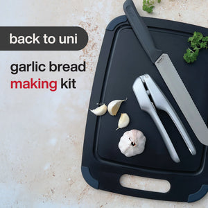 A black tray with a Zyliss bread knife, garlic press, fresh garlic cloves, and parsley. Text reads: Garlic Bread Making Kit. All items are neatly arranged on a light stone background.