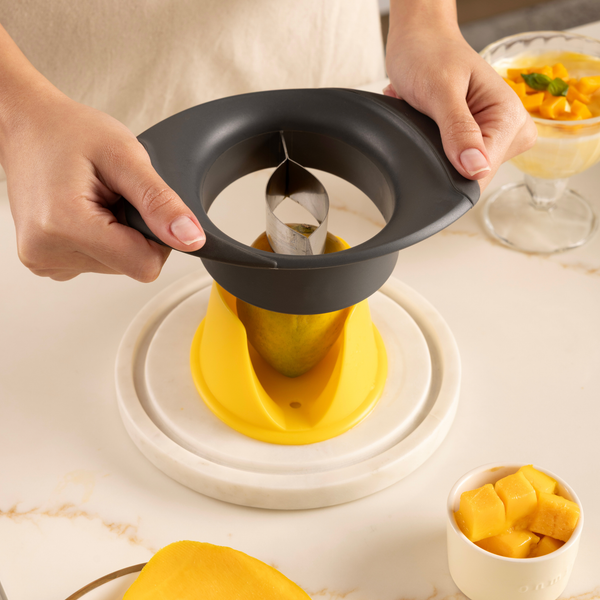 A person uses a Zyliss Mango Splitter to cut a mango on a white countertop, with cubed mango pieces and a dessert in a glass bowl nearby.