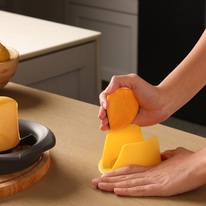 A person uses a yellow Zyliss Mango Splitter to cut a mango over a wooden table, with part of a kitchen counter and another sliced mango visible in the background.