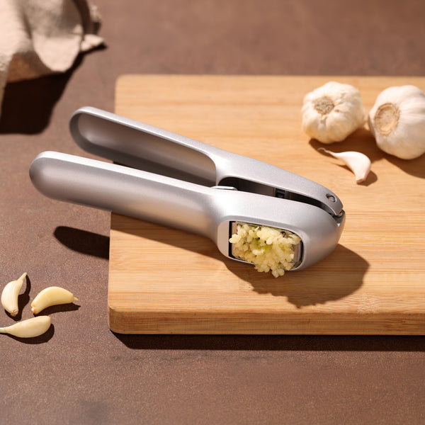 A Zyliss Garlic Bread Making Kit rests on a wooden cutting board, accompanied by freshly pressed garlic, whole bulbs, and scattered cloves on a brown surface. Ideal for crafting homemade spreads or enhancing your favorite garlic bread recipes, this essential kitchen tool pairs perfectly with other utensils like a sturdy bread knife.