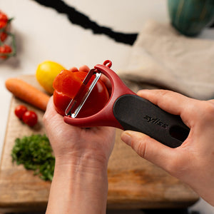 Using the Zyliss Soft Skin Y Peeler, someone peels a tomato over a wooden cutting board surrounded by chopped herbs, carrot, lemon, and cherry tomatoes. This ergonomic tool from Zyliss is ideal for delicate produce.