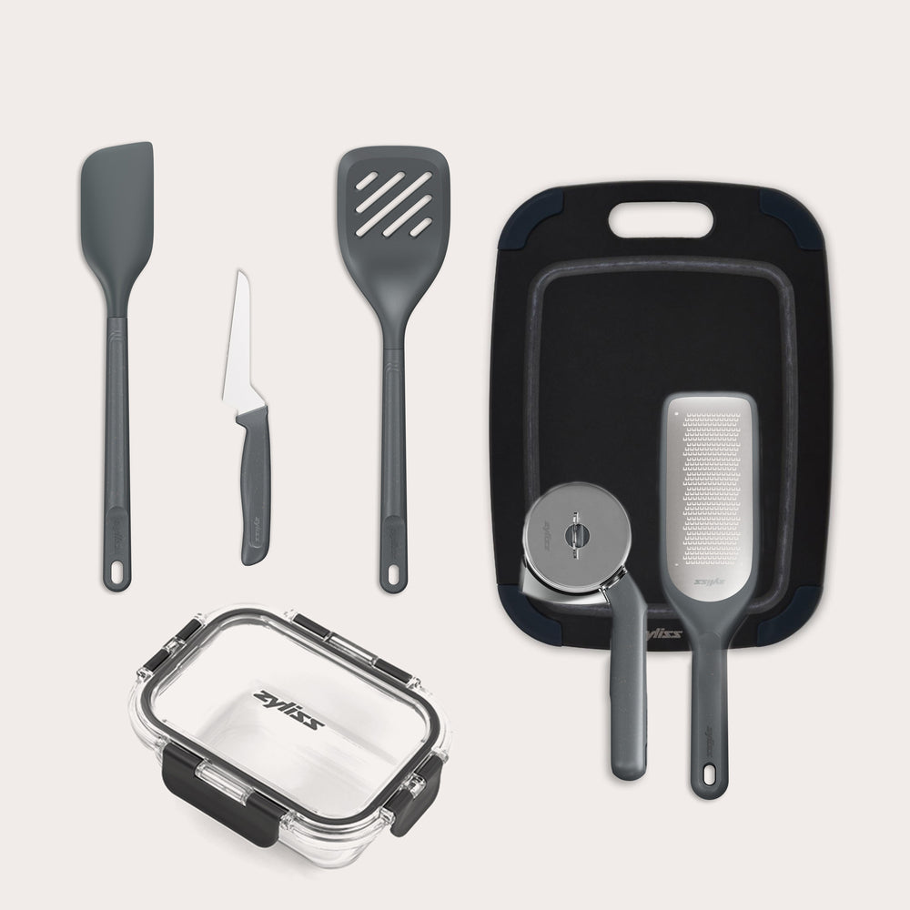 The Zyliss 7-Piece Kitchen Essentials Bundle includes a spatula, slotted turner, paring knife, cutting board, grater, pizza cutter, and a clear rectangular food storage container with a black lid.