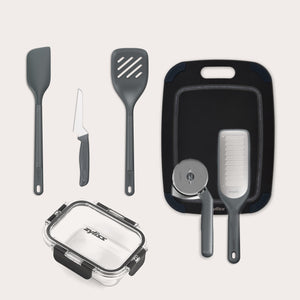 The Zyliss 7-Piece Kitchen Essentials Bundle includes a spatula, slotted turner, paring knife, cutting board, grater, pizza cutter, and a clear rectangular food storage container with a black lid.