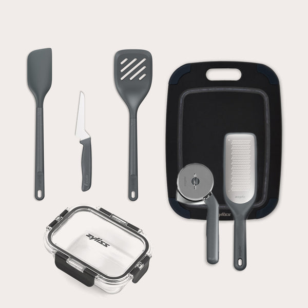 The Zyliss 7-Piece Kitchen Essentials Bundle includes a spatula, slotted turner, paring knife, cutting board, grater, pizza cutter, and a clear rectangular food storage container with a black lid.