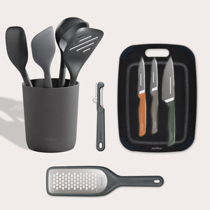 The Zyliss Kitchen Essentials Bundle includes black kitchen tools in a holder, a black peeler, three colorful-handled stainless steel knives with a cutting board, and a flat grater, all displayed on a light background.