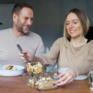 A woman serves a casserole from a Zyliss Glass Container while a man looks on. They sit at a wooden table, sharing meal prep in a cozy indoor setting, highlighting the secure and stylish Zyliss Glass Container. #1.52L/1.61qt