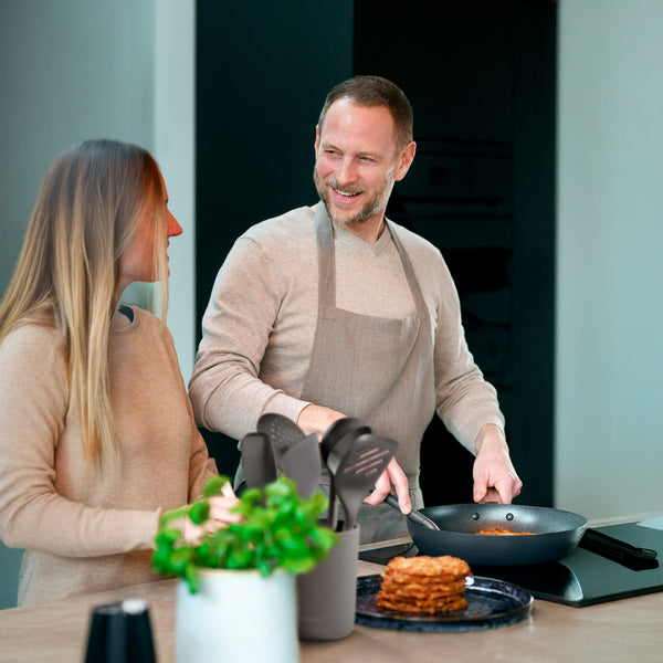 A man in an apron smiles at a woman as he cooks in a modern kitchen. A Zyliss Spoontula rests by pancakes and utensils, while a green potted plant sits in the foreground. #Medium