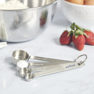 A set of metal measuring spoons rests on a marble countertop near a stainless steel mixing bowl, a bowl of eggs, and several fresh strawberries.