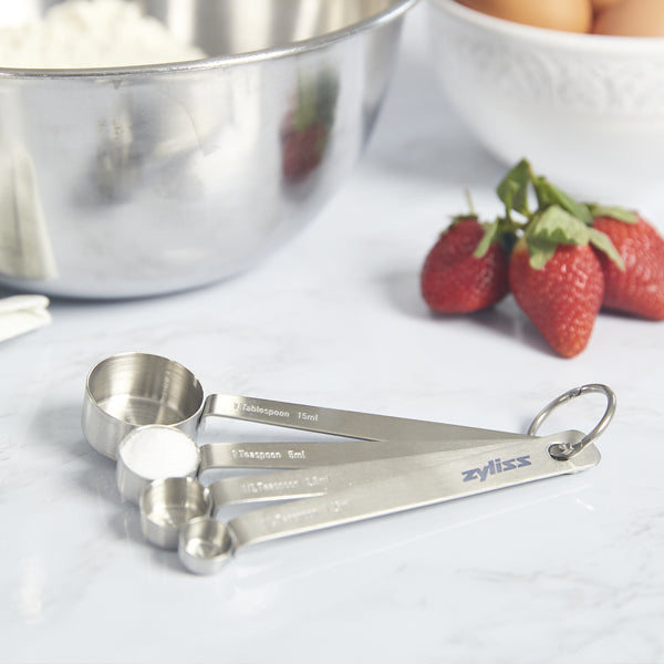 A set of metal measuring spoons rests on a marble countertop near a stainless steel mixing bowl, a bowl of eggs, and several fresh strawberries.