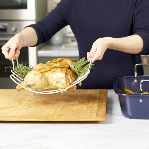 A person places a cooked whole chicken with herbs onto a wooden cutting board using a metal roasting rack in a kitchen setting.