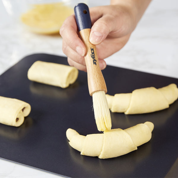 A hand uses a pastry brush to apply egg wash to unbaked croissants and rolls on a black baking tray.
